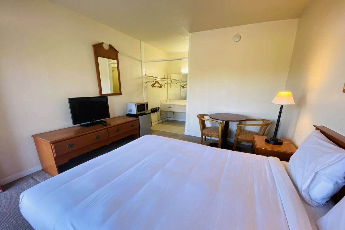 A hotel room with a large bed, a wooden dresser, a television, a microwave, a small table with chairs, and a lamp.