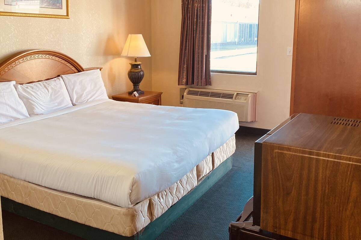 A hotel room with a large bed, a bedside table with a lamp, a window with curtains, and a wooden cabinet.