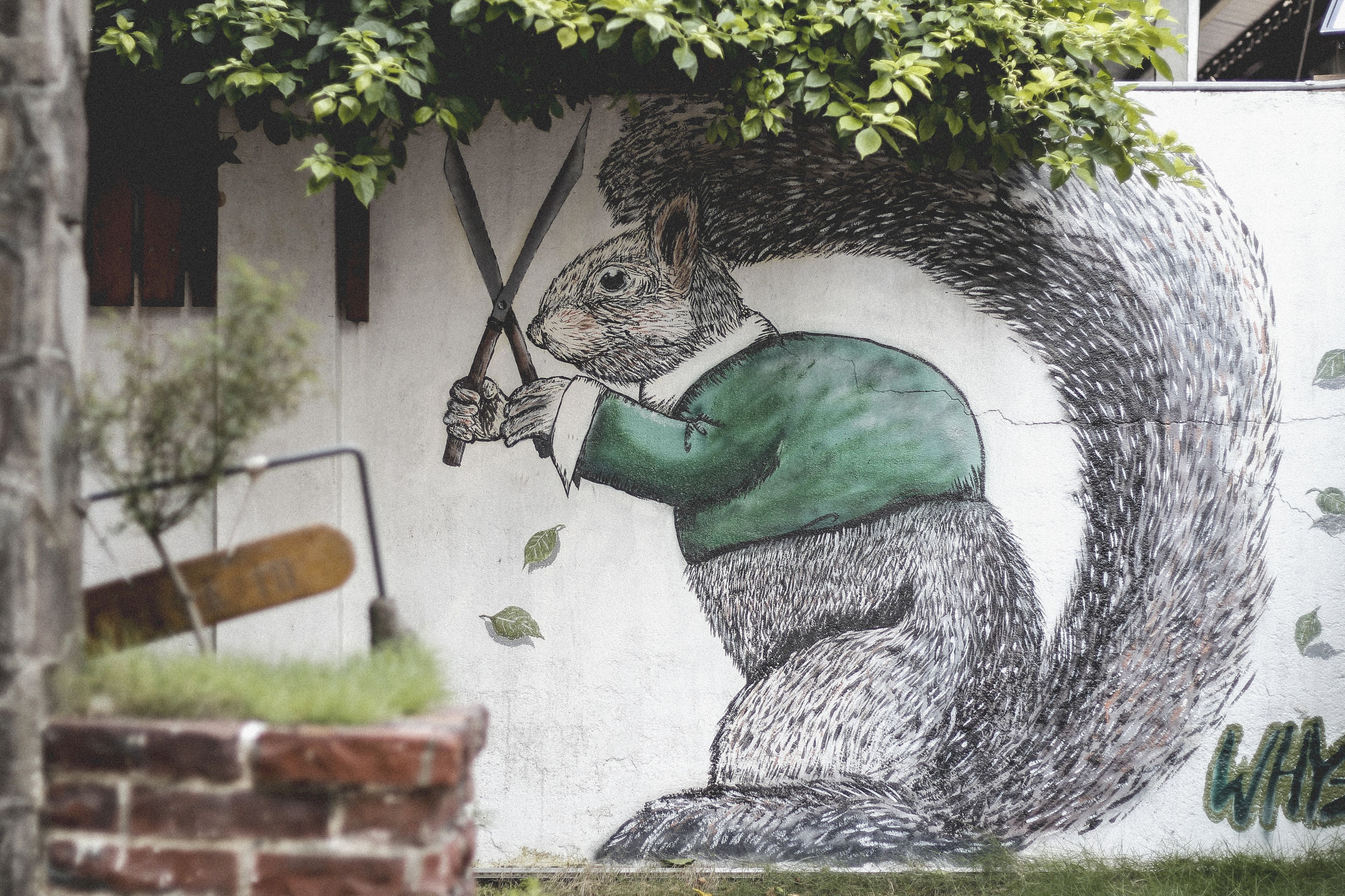 A mural of a squirrel holding a pair of scissors, with a brick wall and a wooden bench in the foreground.