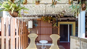 Een overdekt terras met twee gele stoelen van netstof en een kleine ronde tafel, omringd door houten schermen en potplanten.