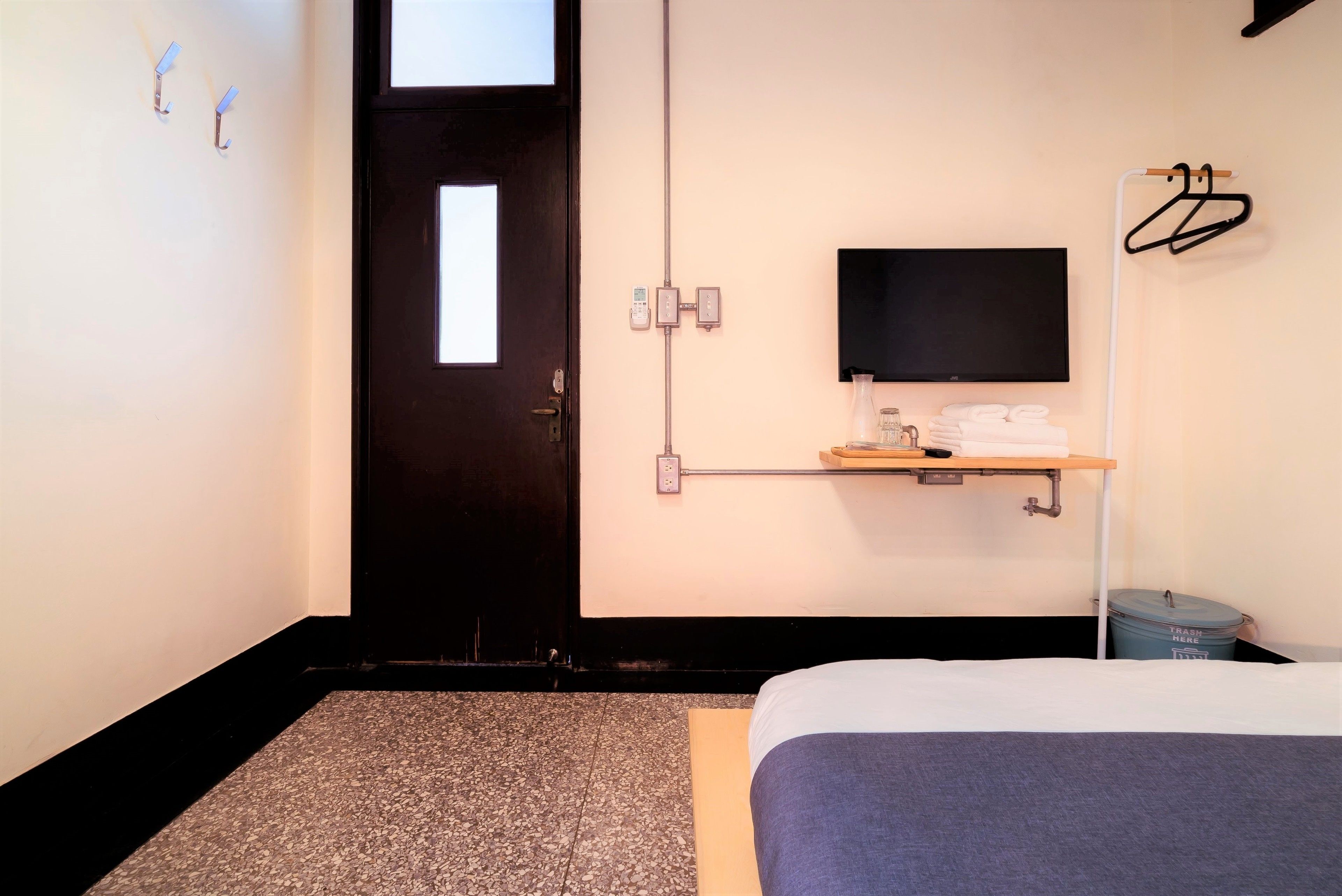 A hotel room with a bed, a TV mounted on the wall, a small shelf with towels, and a trash bin.