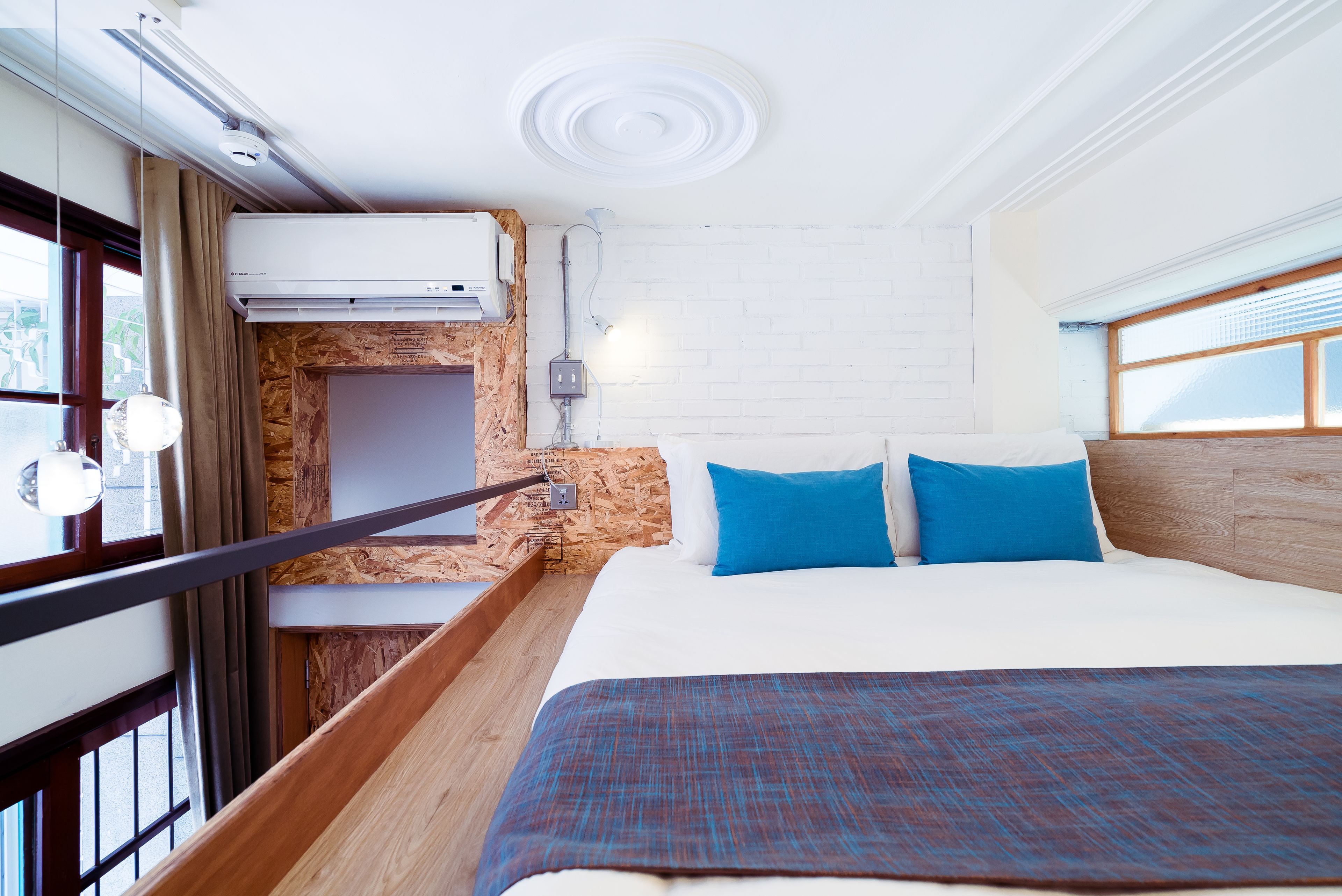 A modern hotel room with a wooden headboard, a large bed with blue pillows, a wooden desk, and a window with curtains.