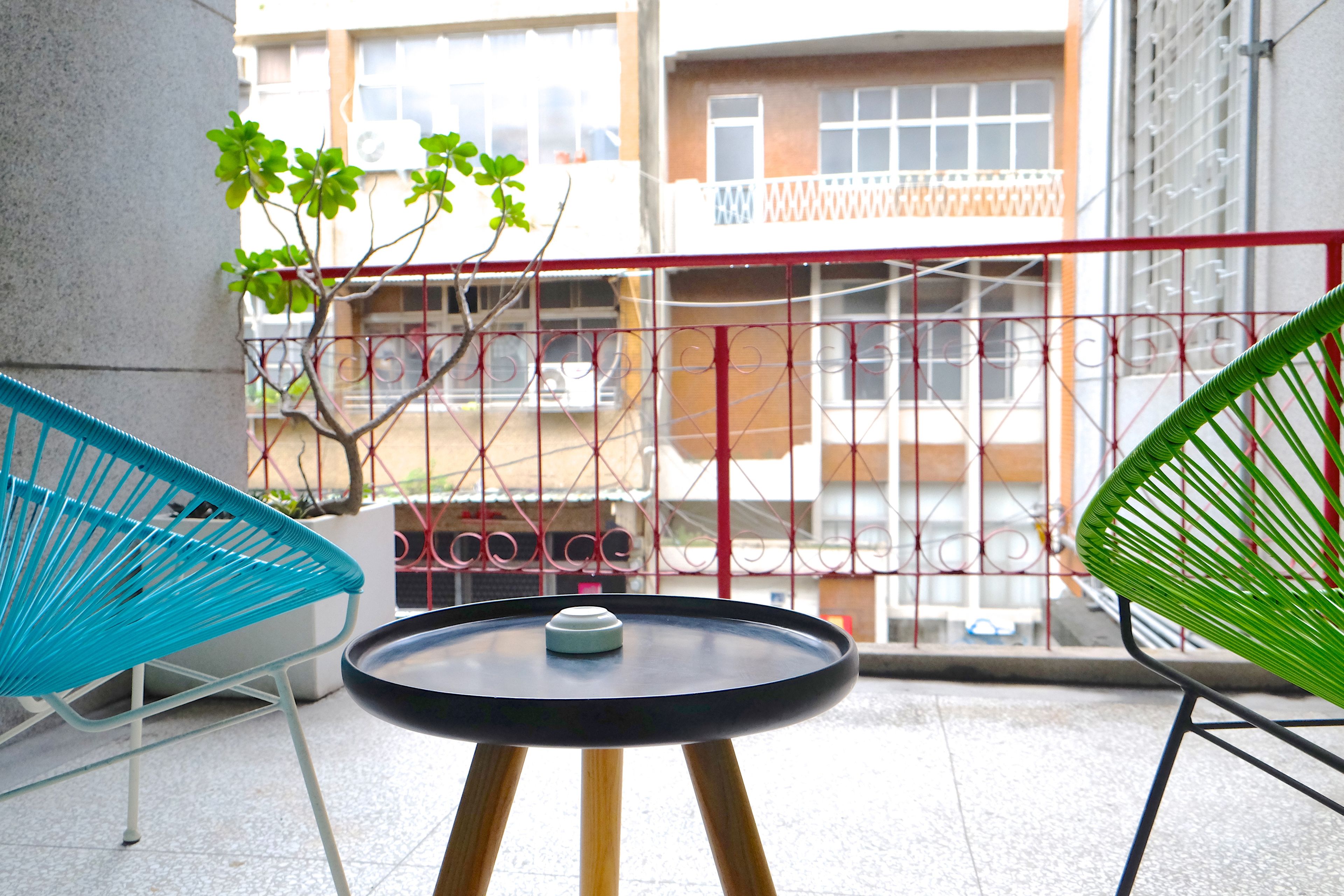 Un balcón con una pequeña mesa redonda y dos sillas, una azul y otra verde, con vistas a una zona residencial.