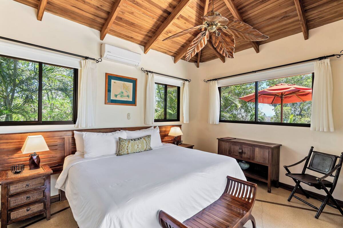 A bedroom with a large bed, wooden headboard, two bedside tables with lamps, a wooden bench, and a ceiling fan.