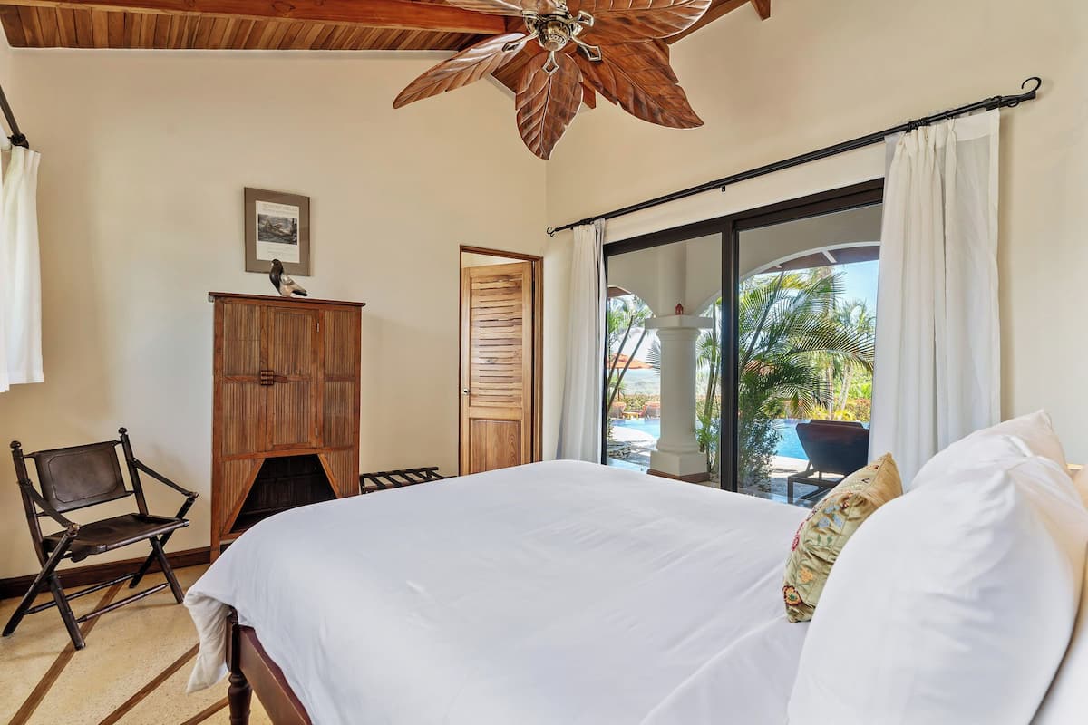 A bedroom with a bed, a wooden wardrobe, a chair, a ceiling fan, and a view of a pool through a glass door.