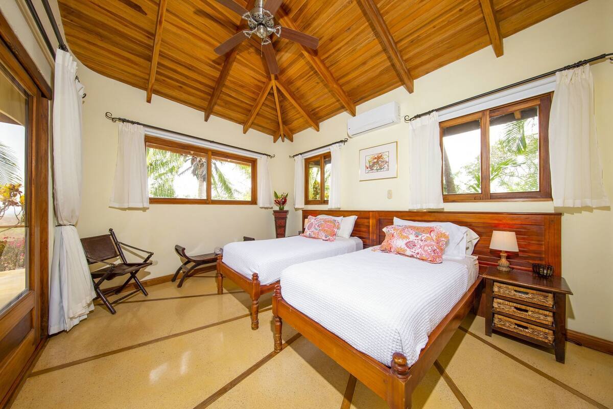 A room with two beds, a ceiling fan, wooden beams, large windows, and a small table with a lamp.