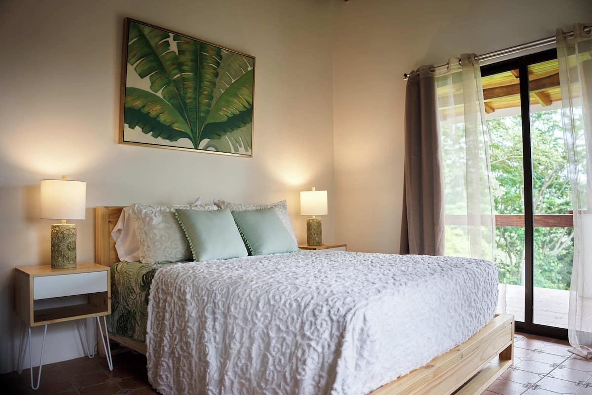 A bedroom with a large bed, two bedside tables with lamps, a wall-mounted painting, and a window with curtains.