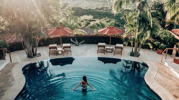 Una piscina con una persona nadando, rodeada de sillas de descanso y sombrillas, con vistas a zonas verdes y colinas al fondo.