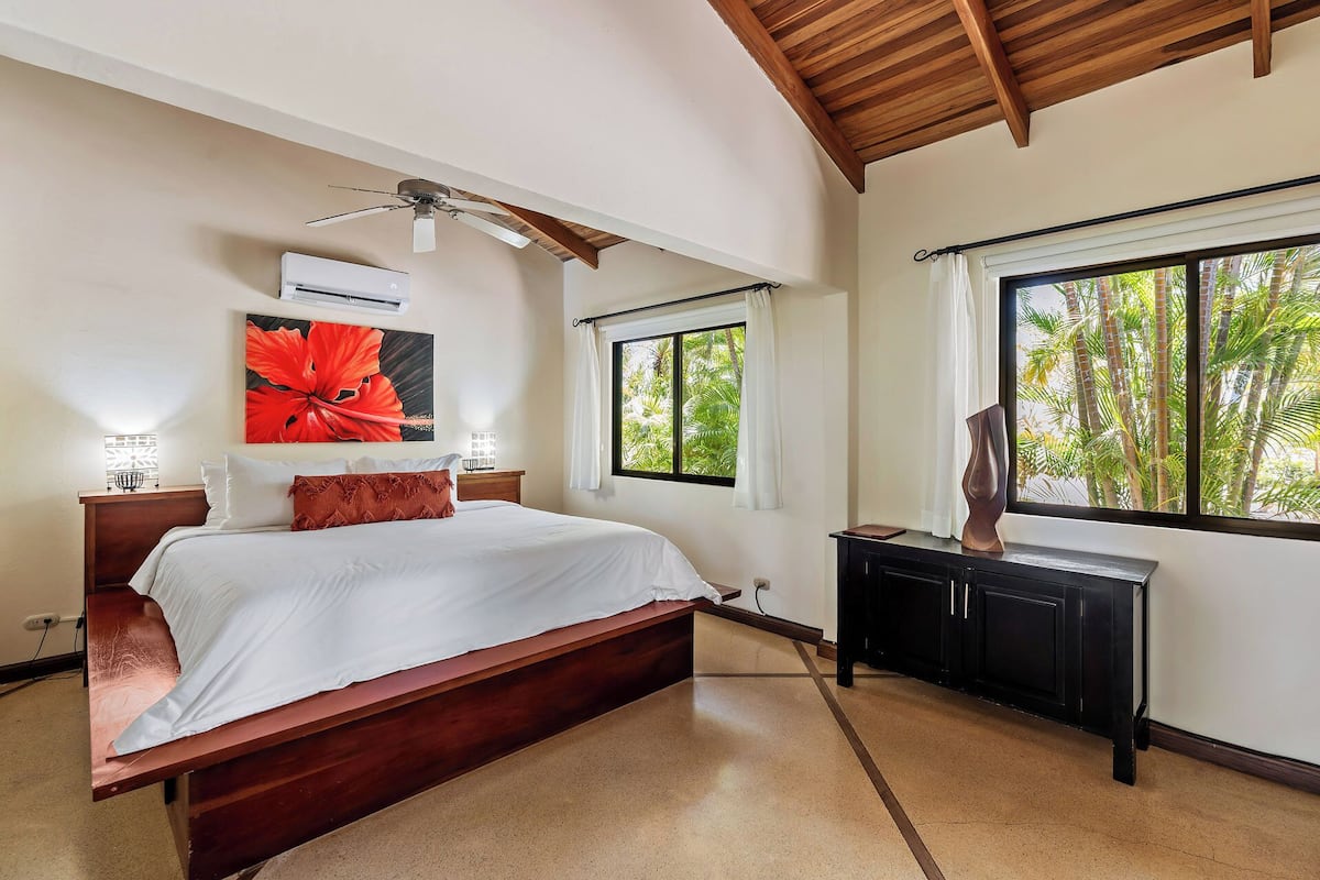 A bedroom with a bed, a ceiling fan, a window with curtains, and a view of greenery outside.