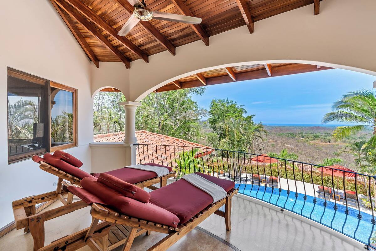 A balcony with two wooden lounge chairs, a ceiling fan, and a view of a swimming pool and landscape.