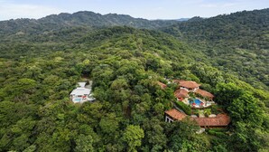 Un resort con varias edificaciones rodeado de un bosque denso, que cuenta con una piscina y una cancha de tenis.