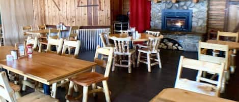 A rustic wooden interior with tables and chairs, a stone fireplace, and a red curtain.