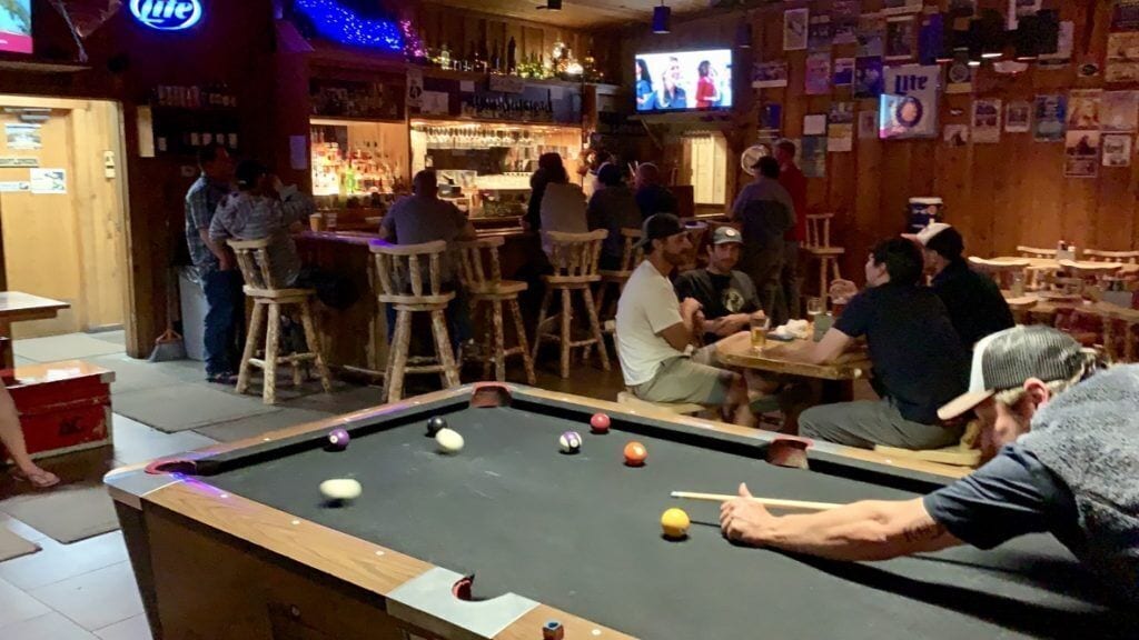 A pool table in a bar with stools around it.