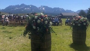 An outdoor seating area with large flower arrangements in barrels, a clear sky, and mountains in the background. - Mountain Village Lodge (Stanley)