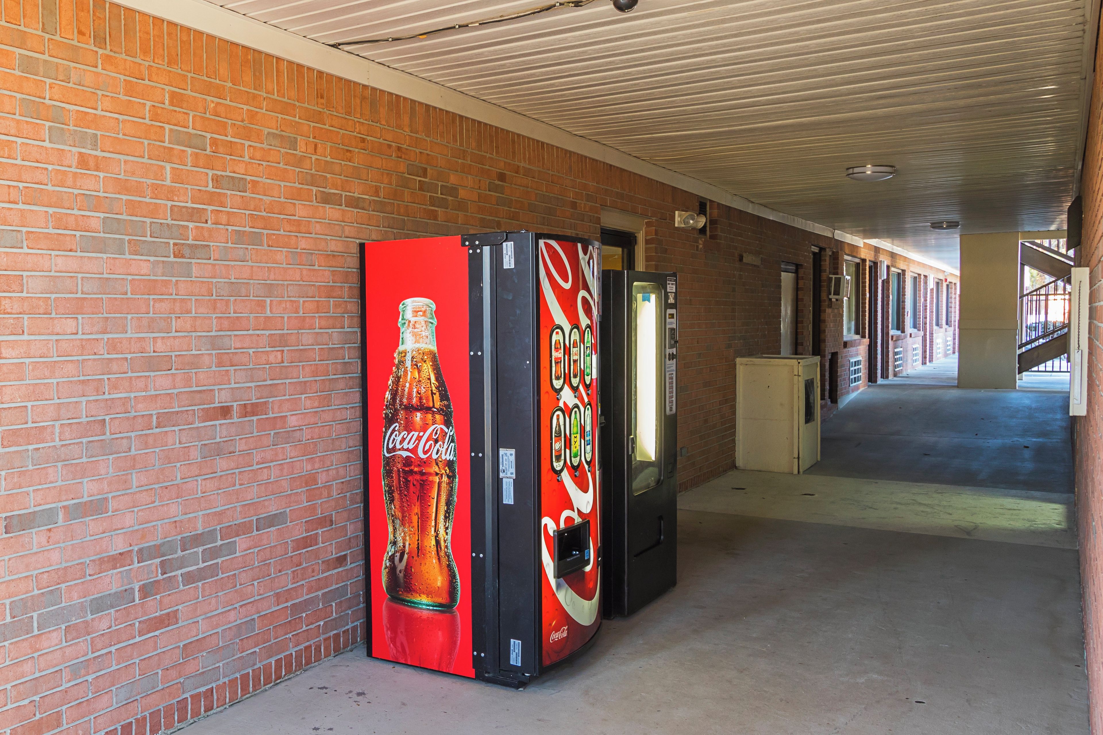 A corridor with brick walls and a ceiling with a linear pattern. There are vending machines with Coca-Cola branding on the wall.