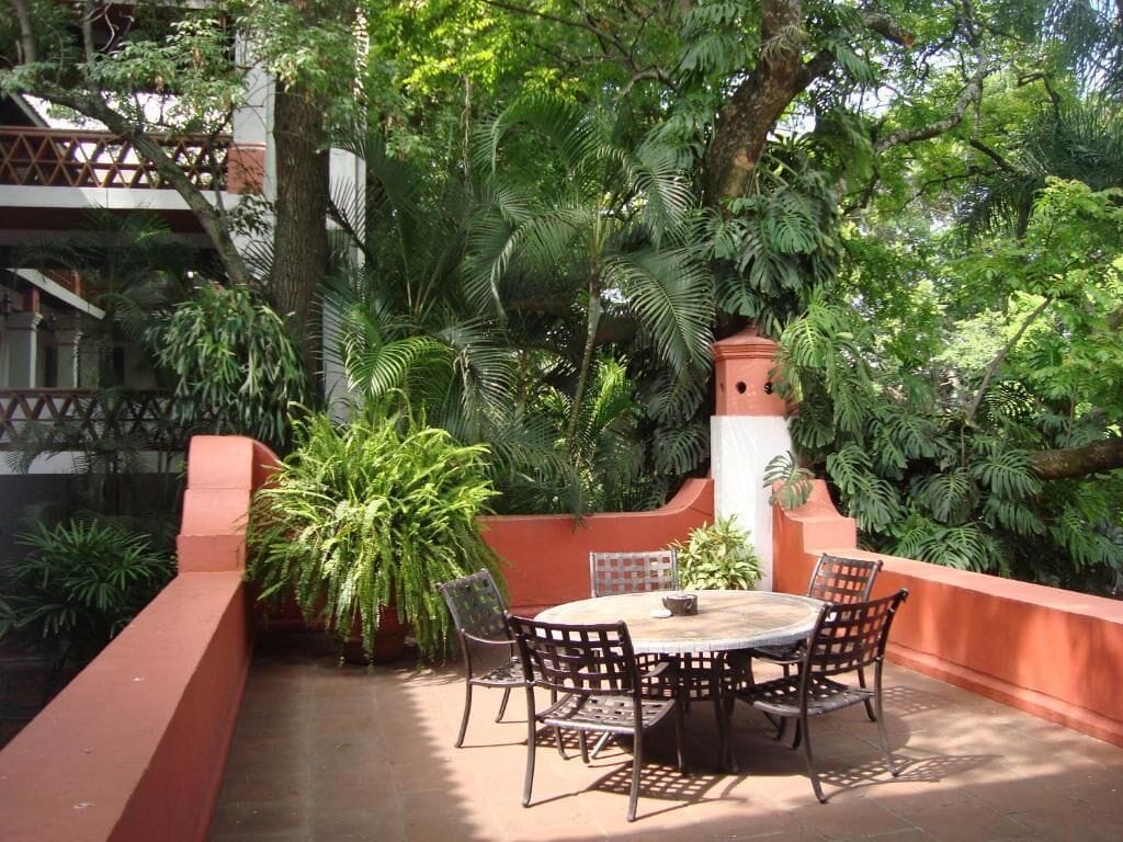 An outdoor patio with a round table and chairs, surrounded by lush greenery and a red wall.