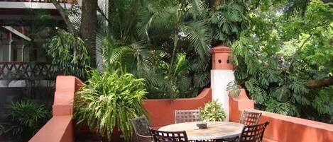 An outdoor patio with a round table and chairs, surrounded by lush greenery and a red wall.