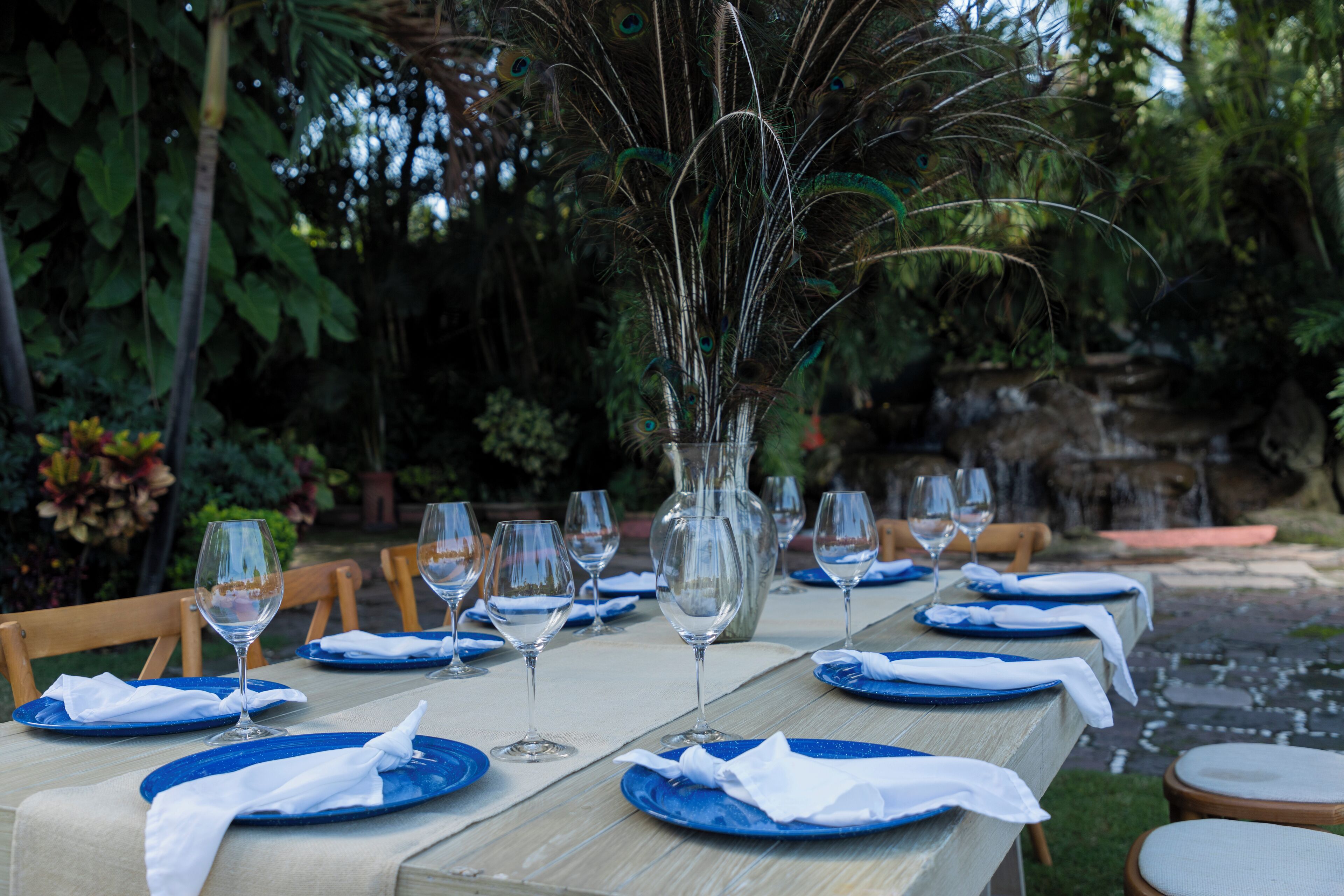 Une table à manger dressée avec des assiettes bleues, des serviettes blanches et des verres à vin, entourée de chaises et agrémentée d’un vase décoratif.