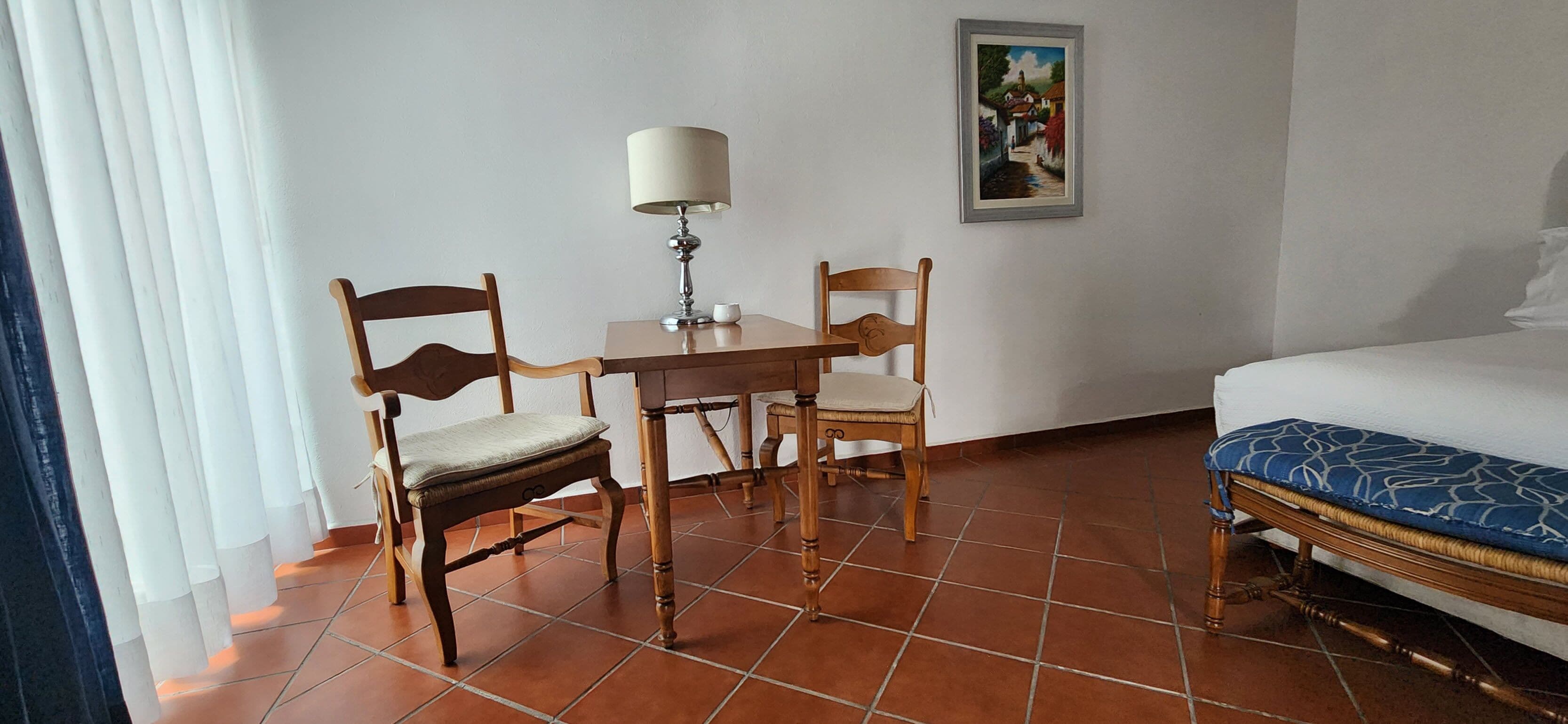 Een hotelkamer met een bed, twee houten stoelen, een bijzettafeltje met een lamp, een schilderij aan de muur en een raam met jaloezieën.