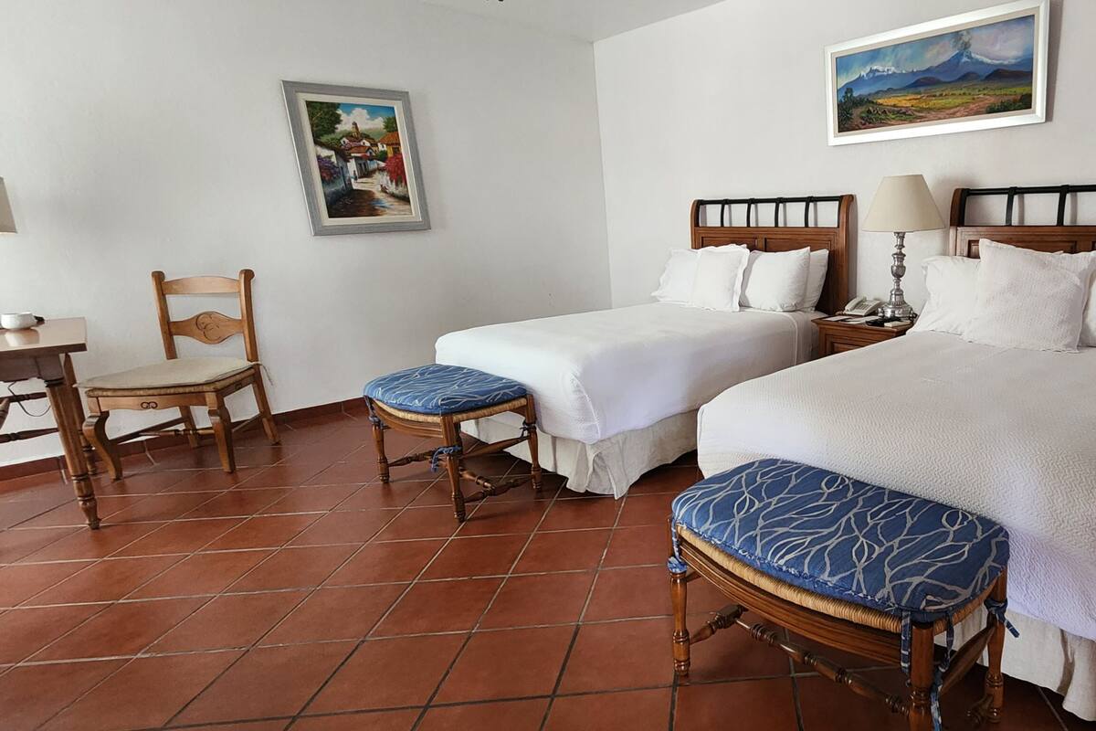 A hotel room with two beds, a desk, chairs, and two paintings on the walls.