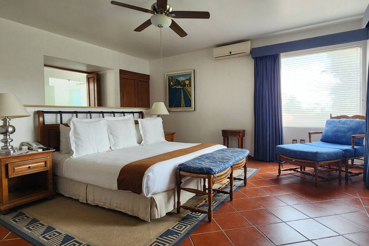 A hotel room with a bed, bedside tables, a desk, a chair, a ceiling fan, and a window with curtains.