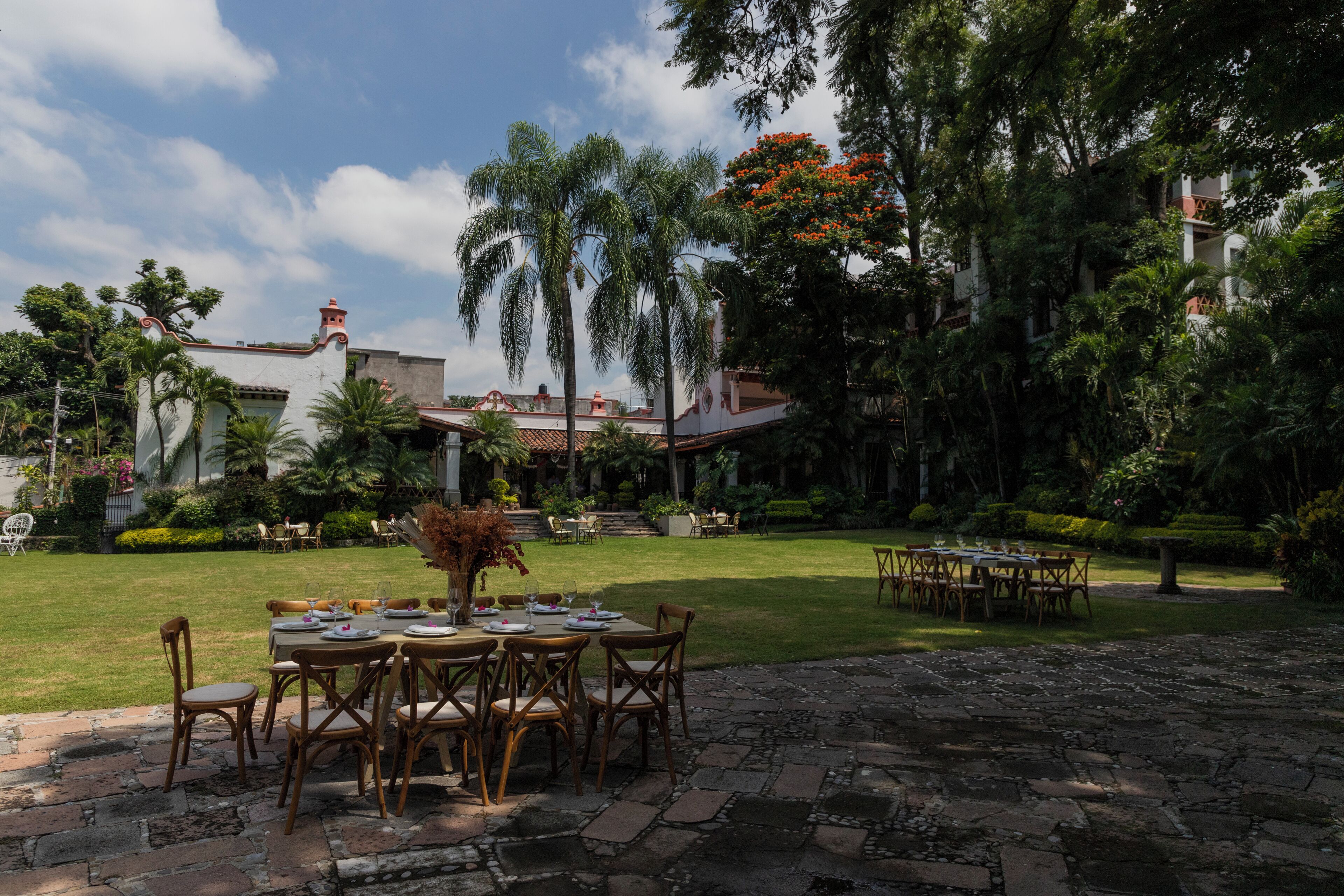 An outdoor dining area with tables and chairs, surrounded by lush greenery and a building with a red roof.