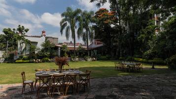 An outdoor dining area with tables and chairs, surrounded by lush greenery and a building with a red roof.