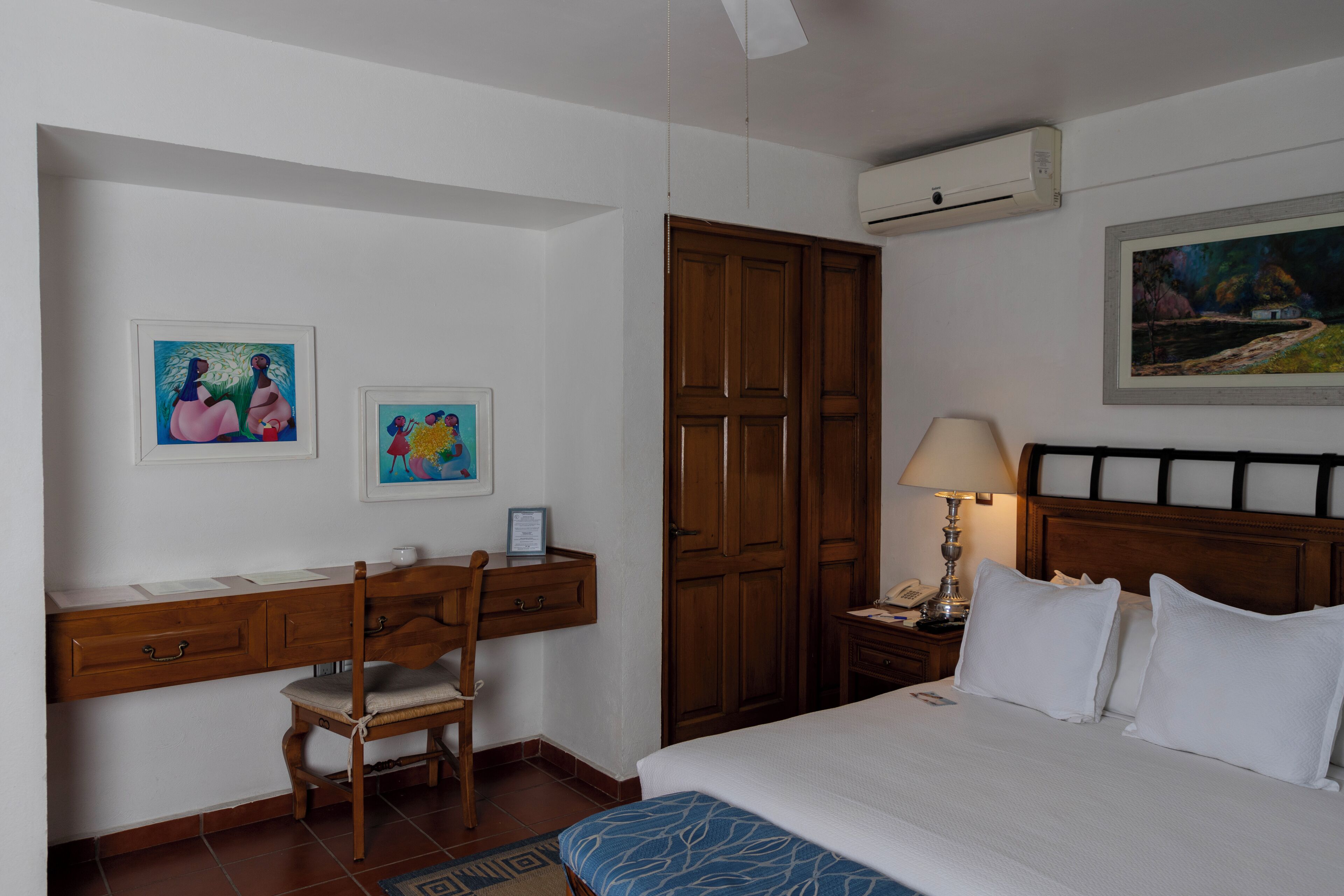 Una habitación de hotel con cama, escritorio con silla, armario, mesita de noche con lámpara y dos cuadros enmarcados en la pared.