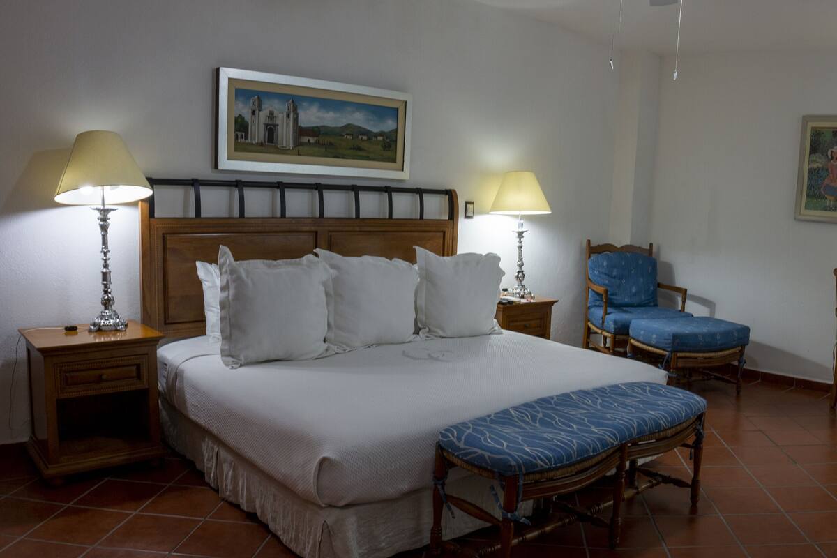 A hotel room with a large bed, two bedside tables with lamps, a wooden headboard, a blue chair, a wooden chair, a small table, and a painting on the wall.