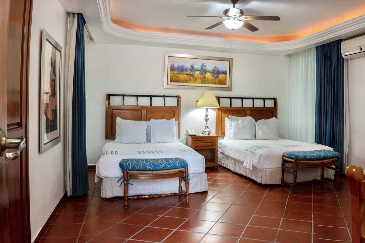 A hotel room with two beds, a ceiling fan, a painting, and a nightstand with a lamp.