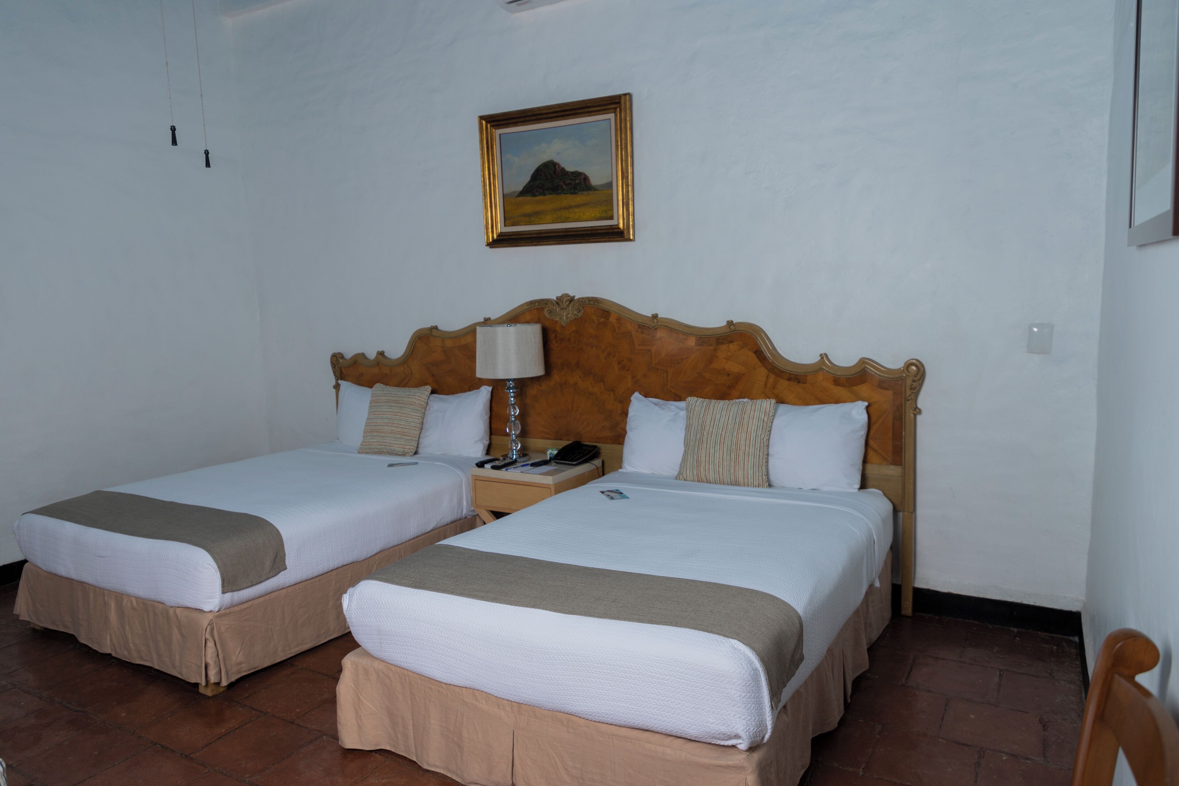 A hotel room with two single beds, a wooden headboard, a nightstand with a lamp, and a framed picture on the wall.
