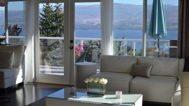 A spacious living room with a white sofa, a glass coffee table, and a view of a lake and mountains through large windows. - Le Chateau du Lac (West Kelowna)
