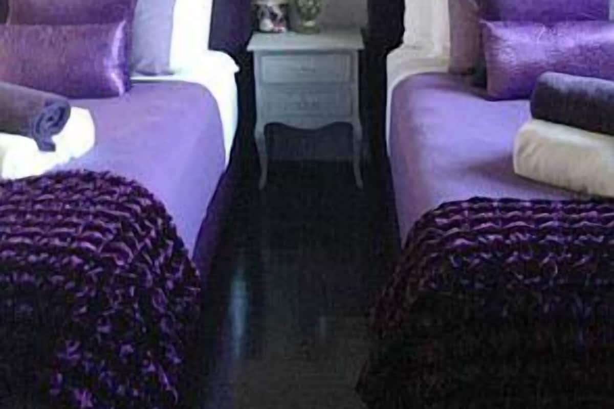 Habitación con dos camas, cada una con ropa de cama y almohadas moradas, una mesita de noche pequeña con una lámpara, y un cartel en la pared que dice 
