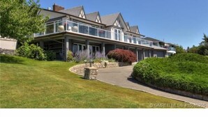 A large, two-story house with a balcony on the upper floor, surrounded by well-maintained lawns and shrubs. - Le Chateau du Lac (West Kelowna)