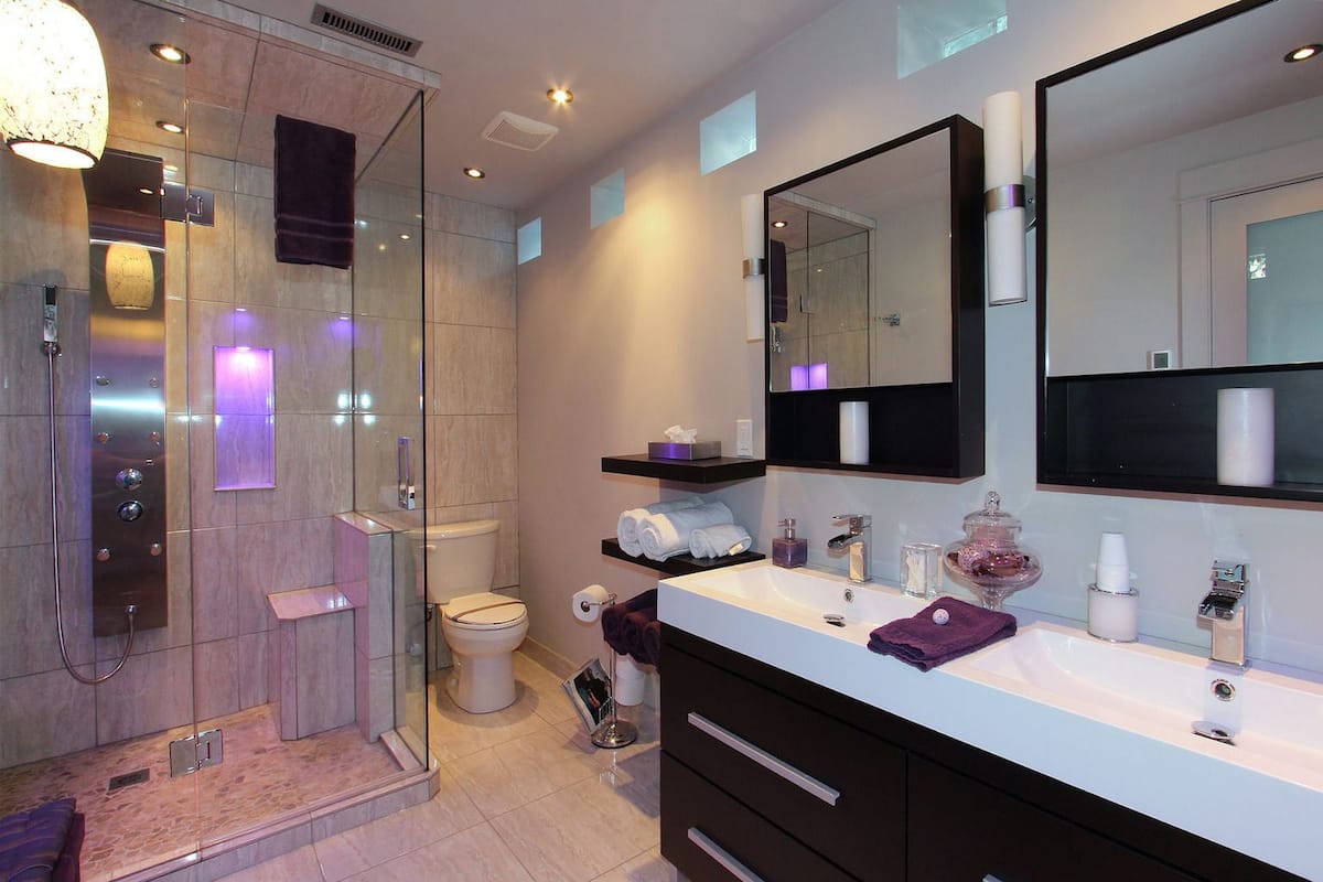 Un baño moderno con ducha acristalada, inodoro, tocador con un espejo grande y lavamanos con diversos artículos de aseo.