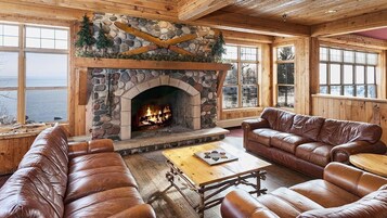 A cozy cabin interior with a stone fireplace, leather furniture, and large windows overlooking a body of water.