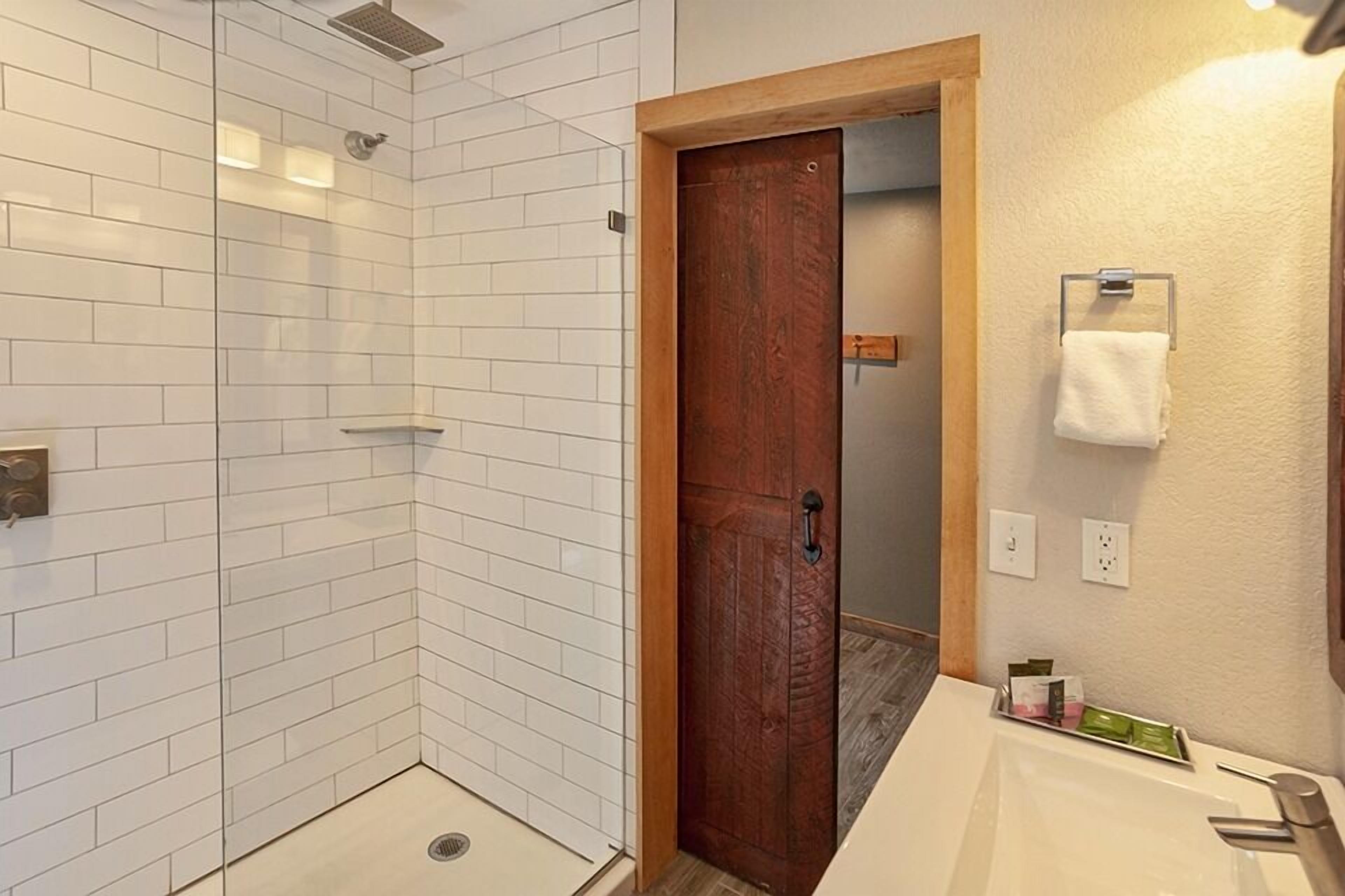 Un baño con paredes de azulejos blancos, una puerta de madera, una ducha y un lavamanos con artículos de aseo.