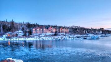 A large lodge with multiple buildings by a frozen river, surrounded by snow-covered trees.