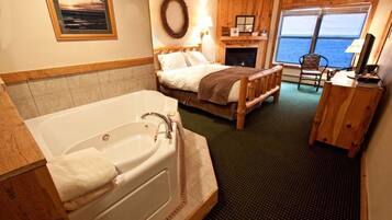 A room with a large bathtub, a bed with a wooden headboard, a fireplace, and a view of the sea through a window.