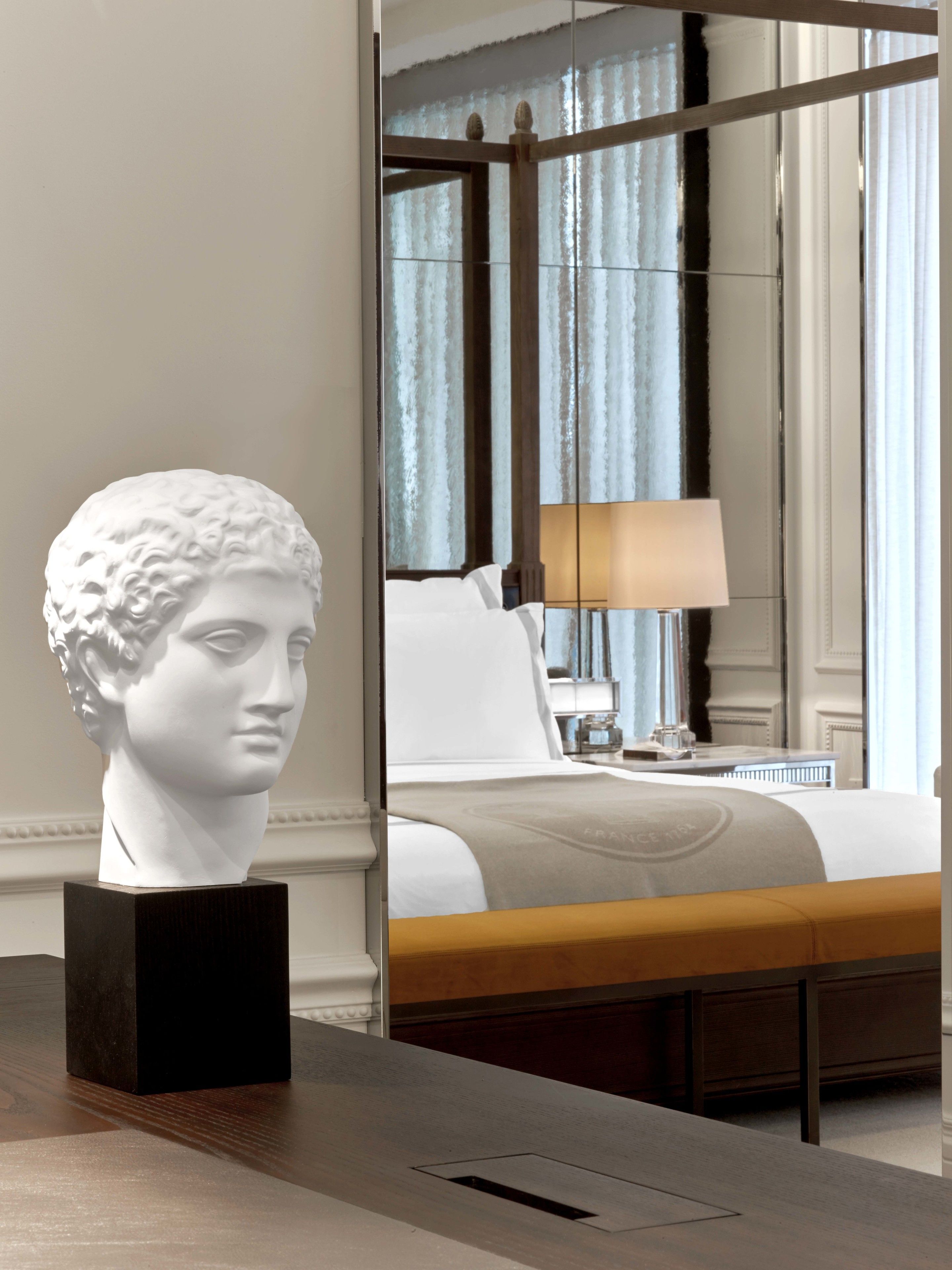 A white bust on a dark wooden table in a hotel room with a bed, a lamp, and a large mirror reflecting the room.