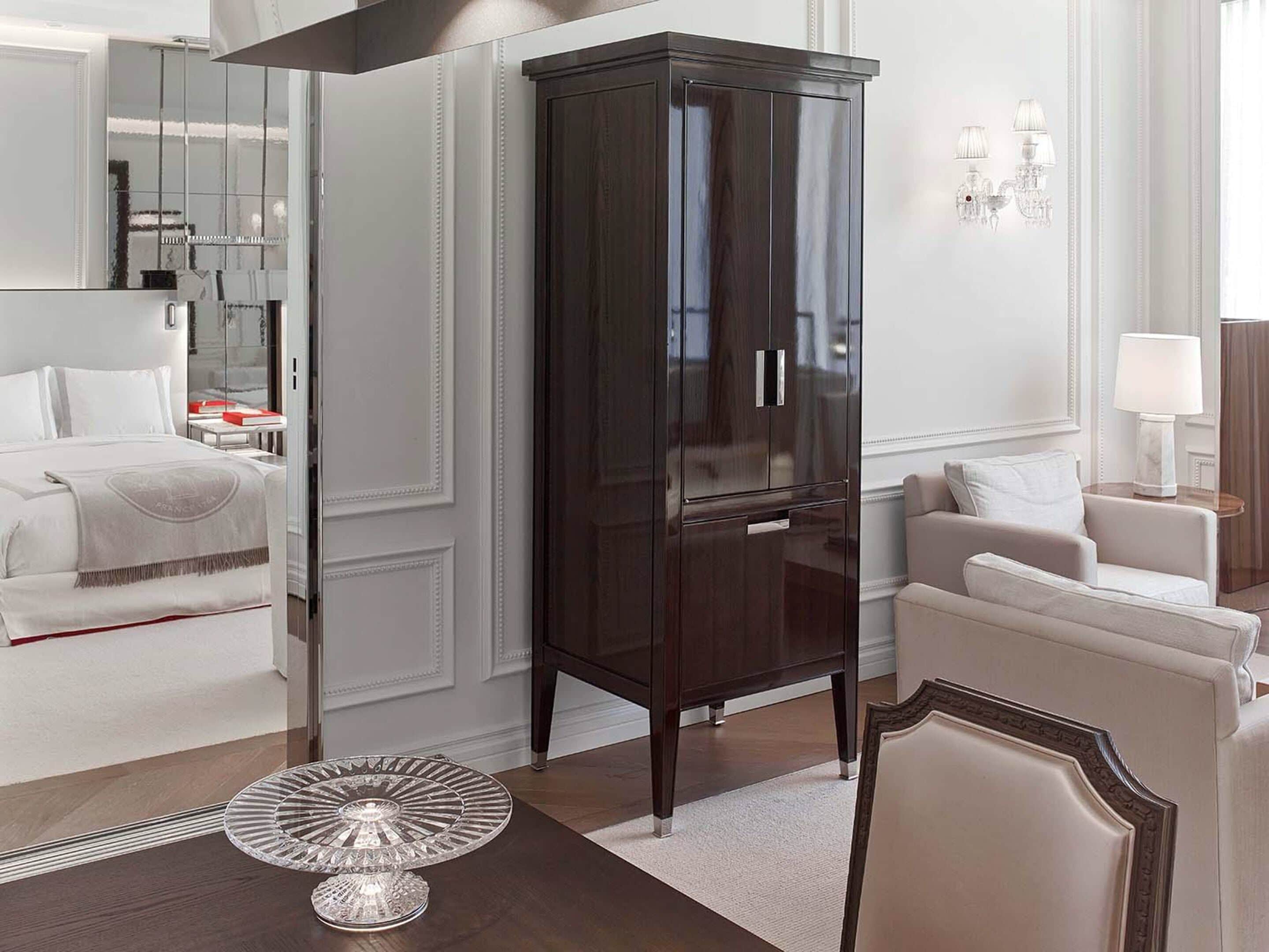 A modern hotel room with a large dark wooden wardrobe, a bed with white linens, a white armchair, and a small crystal table.