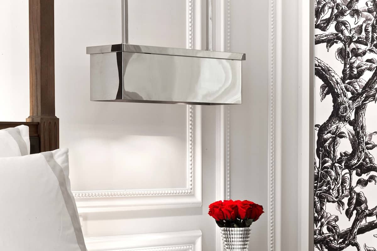 Habitación de hotel ordenada con una cama, una mesita de noche con un jarrón de rosas rojas, un libro rojo y una lámpara colgante moderna.