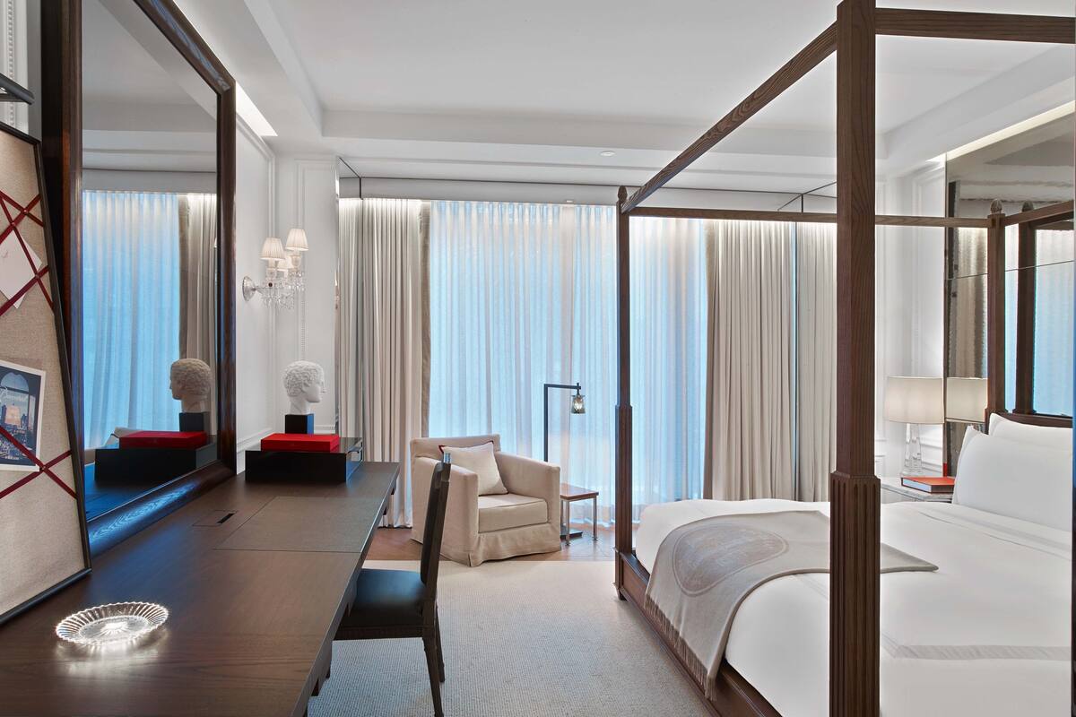 Amplia habitación de hotel con una cama grande, un lecho con dosel de madera, un escritorio con silla, una mesita de noche con lámpara y un espejo grande.