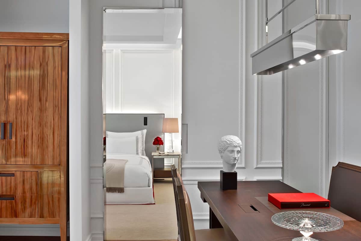 Habitación de hotel con un armario de madera, una cama con ropa blanca, una mesita de noche con una lámpara, una escultura de busto y una caja roja sobre una mesa.