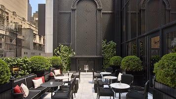 An outdoor seating area with tables and chairs, surrounded by greenery and a building with a patterned facade.