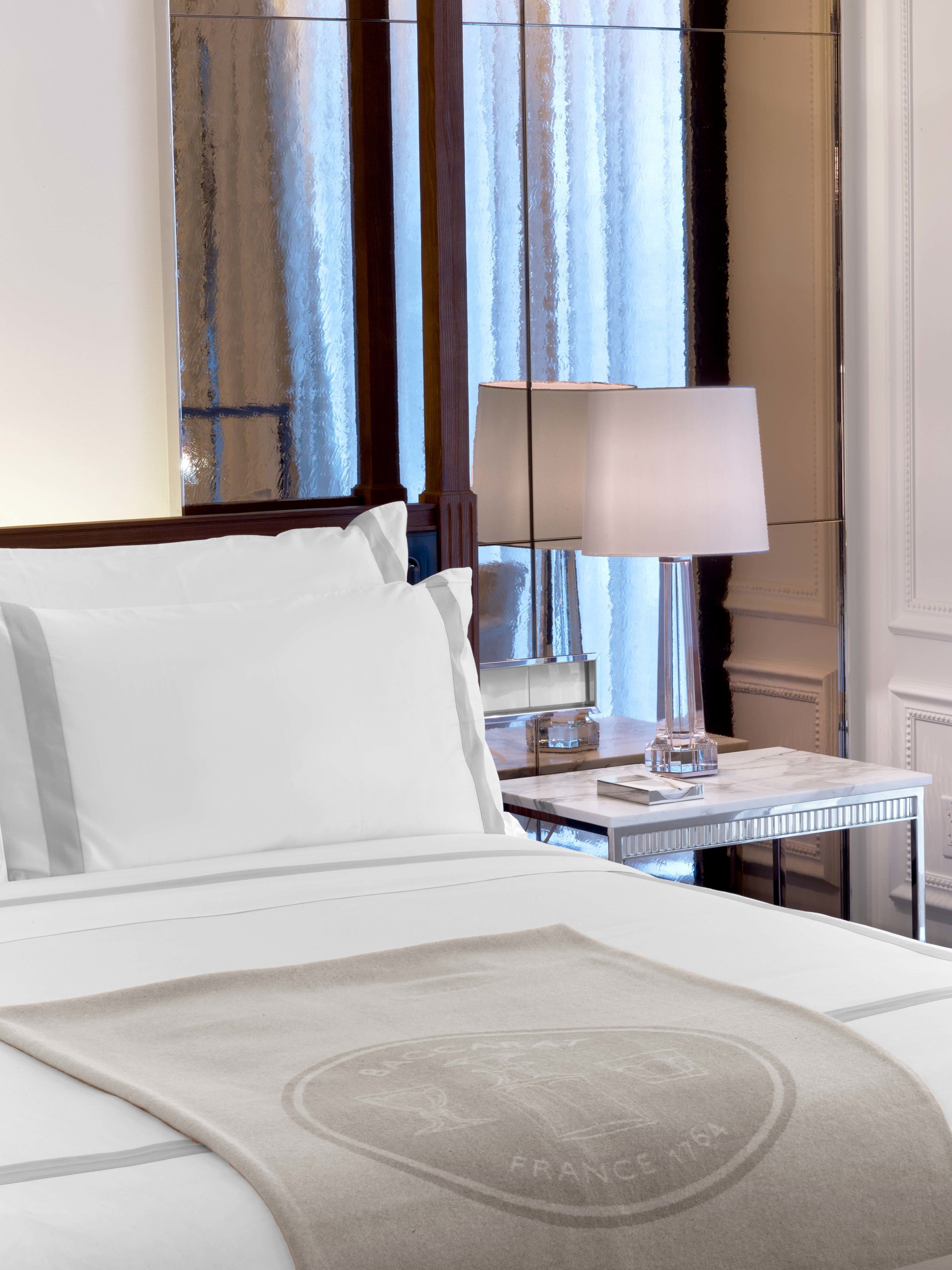 A hotel room with a bed, a nightstand, a lamp, and a view of the outside through curtains.