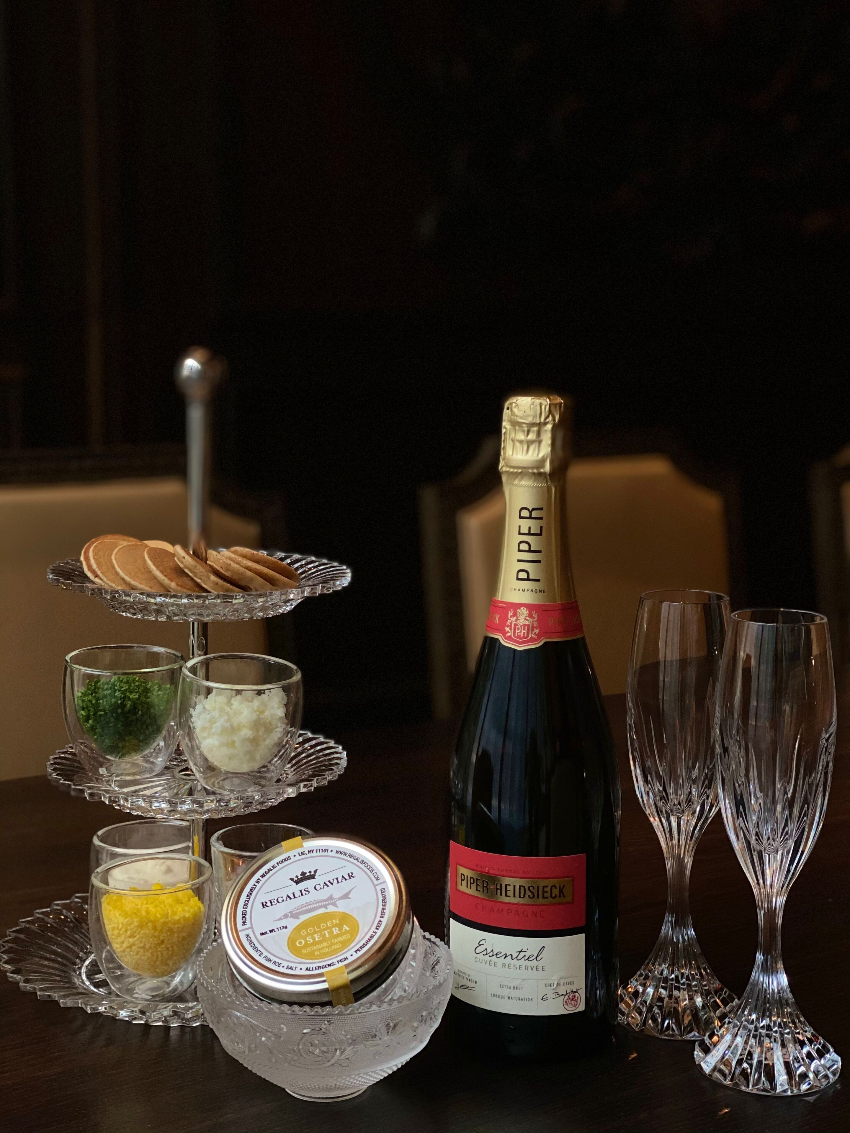 A table with a bottle of PIPER HEIDSIECKER champagne, two glasses, a tiered serving tray with food items, and a container of golden osetra caviar.