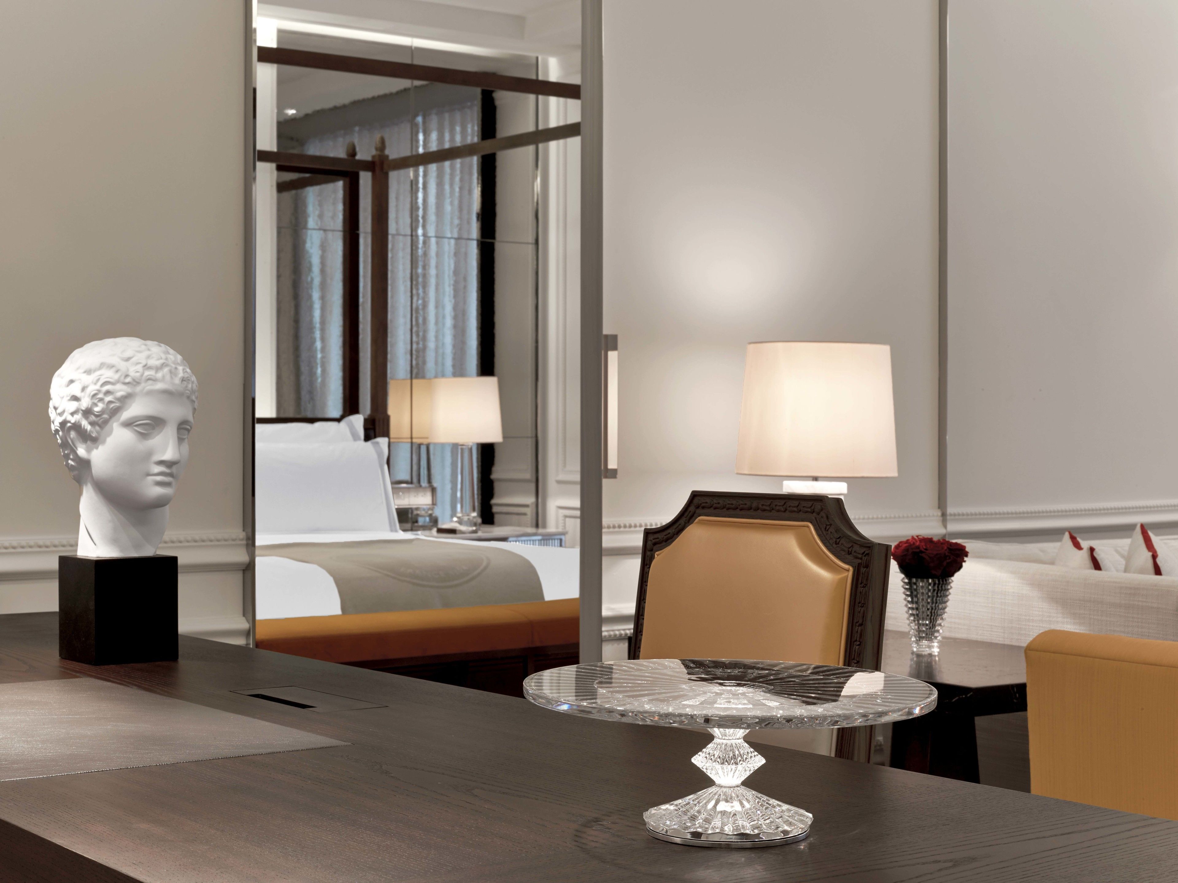 A hotel room with a bed, a nightstand with a lamp, a vase with a flower, and a bust on a table.