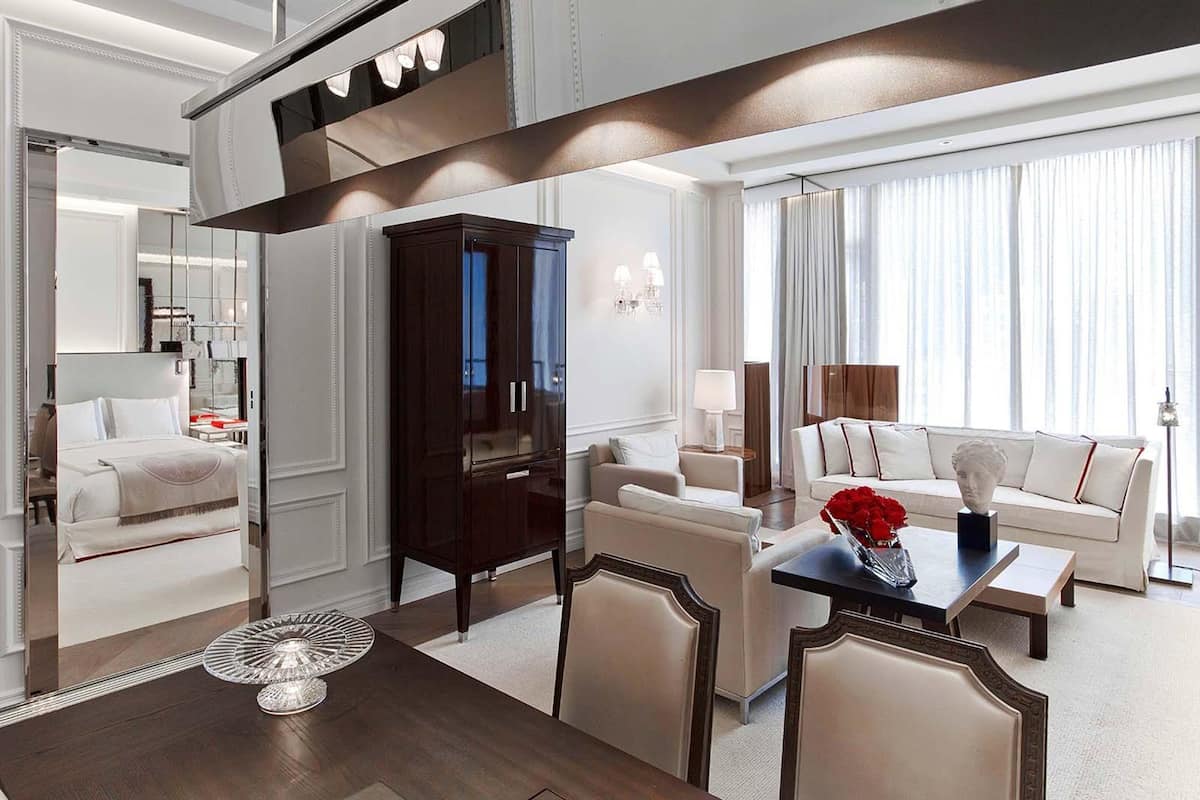Una habitación de hotel moderna con zona de comedor, un mueble grande de madera oscura, un sofá blanco y una mesa de comedor con un jarrón de rosas rojas.