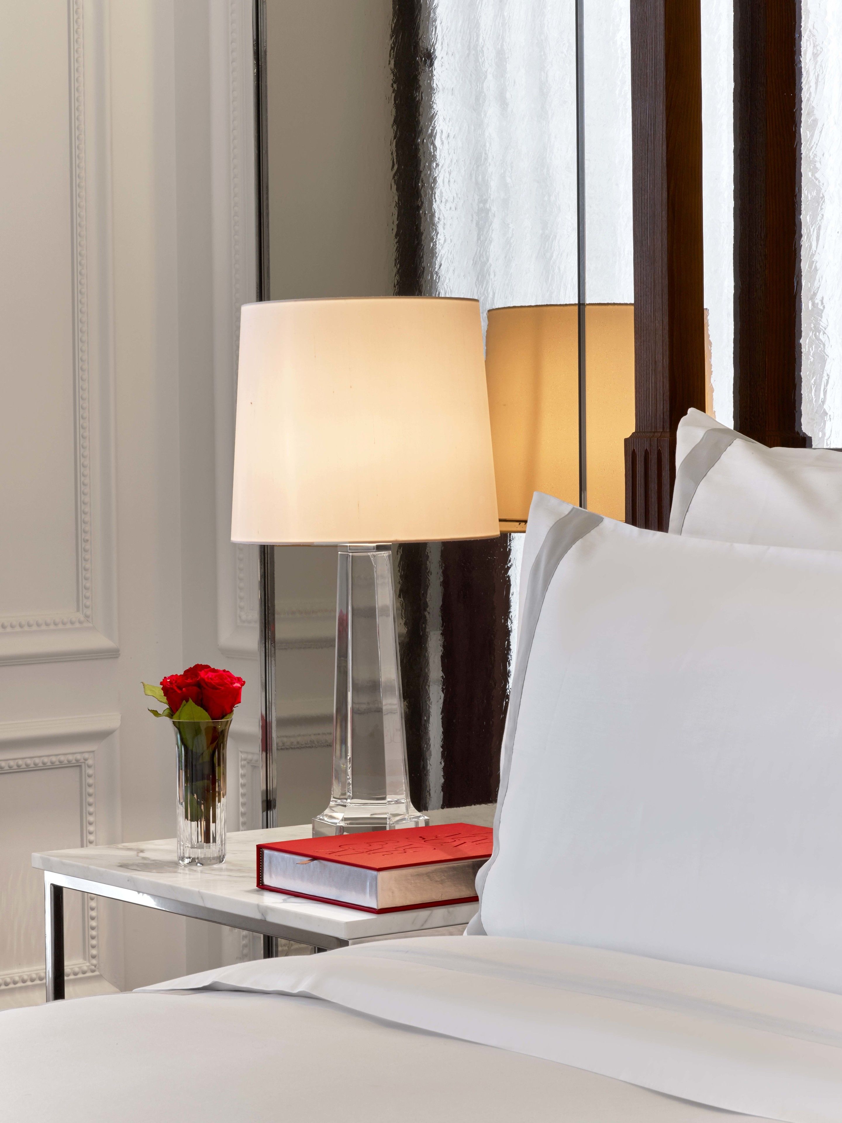Una cama bien hecha con sábanas blancas, una mesita de noche con lámpara, un libro y un jarrón con rosas rojas.